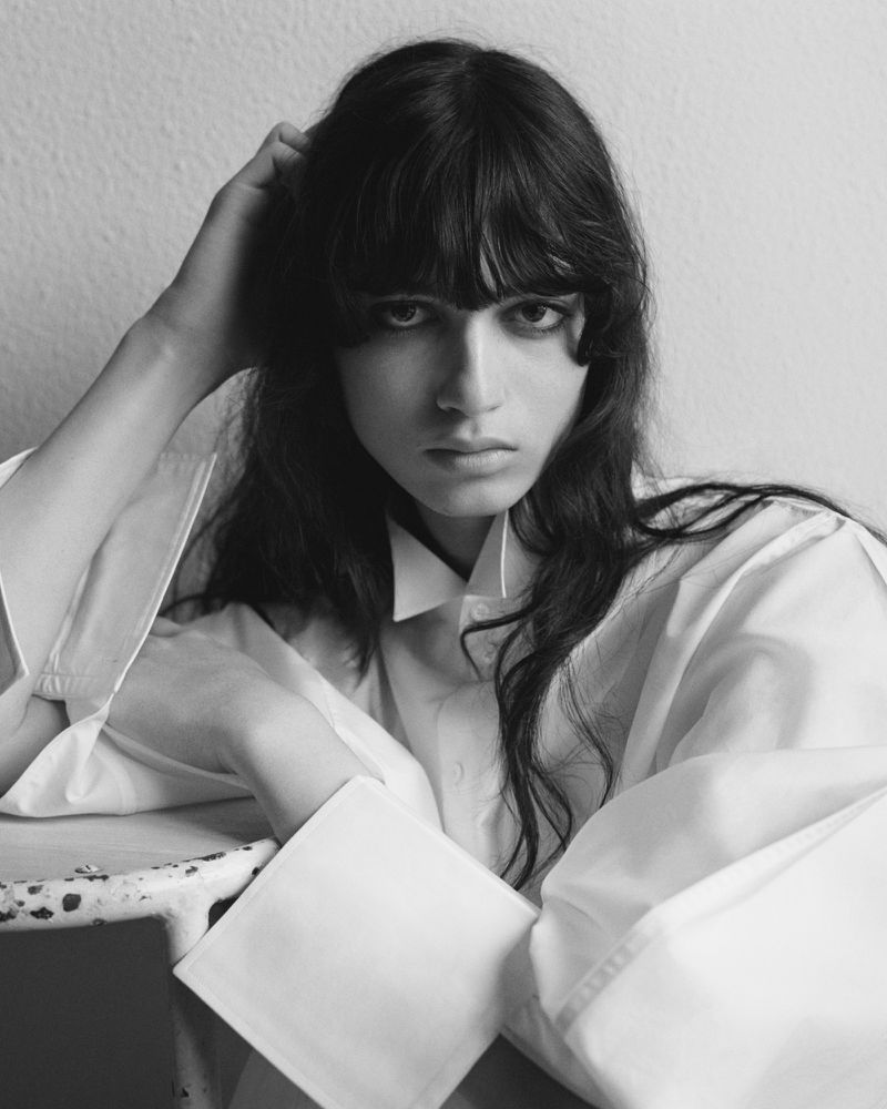 Black and white portrait of a model with long dark hair and bangs wearing an oversized white shirt with wide cuffs.