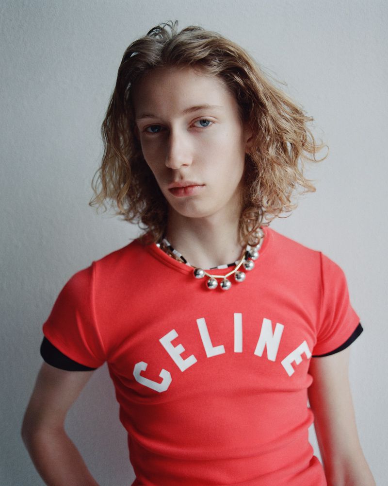 Portrait of a young adult with blonde curly hair and a red Celine t-shirt, posing in front of a neutral grey wall indoors.