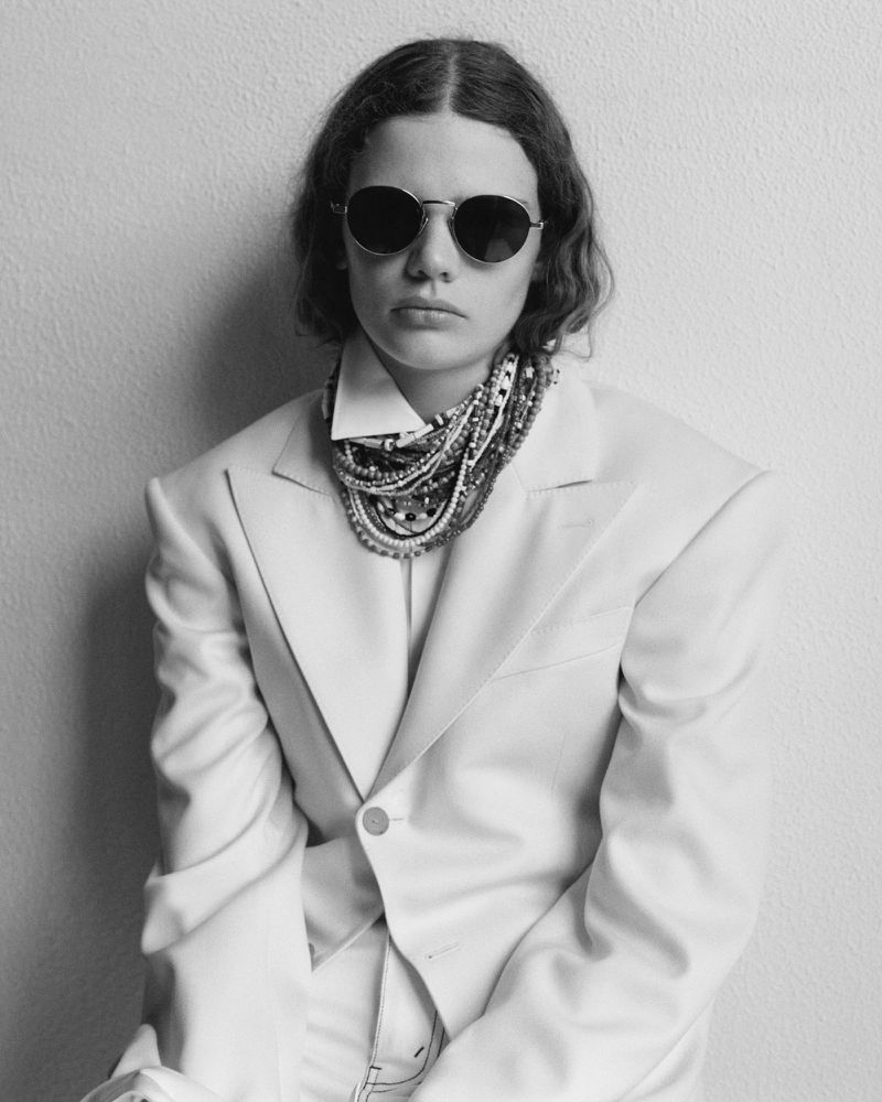 Model wearing dark round sunglasses, an oversized tailored blazer, and a multi-strand beaded necklace in a black and white portrait.