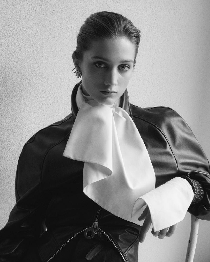 Portrait of a woman in a black jacket and white bow tie, facing forward in a studio with diffused lighting. Black and white image.