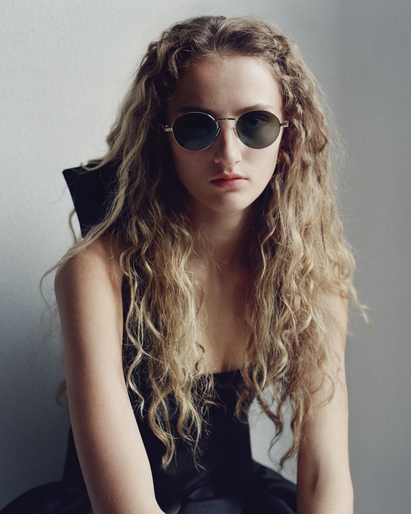 Model with long wavy hair wearing round wire-frame sunglasses and a black garment against a pale neutral background.