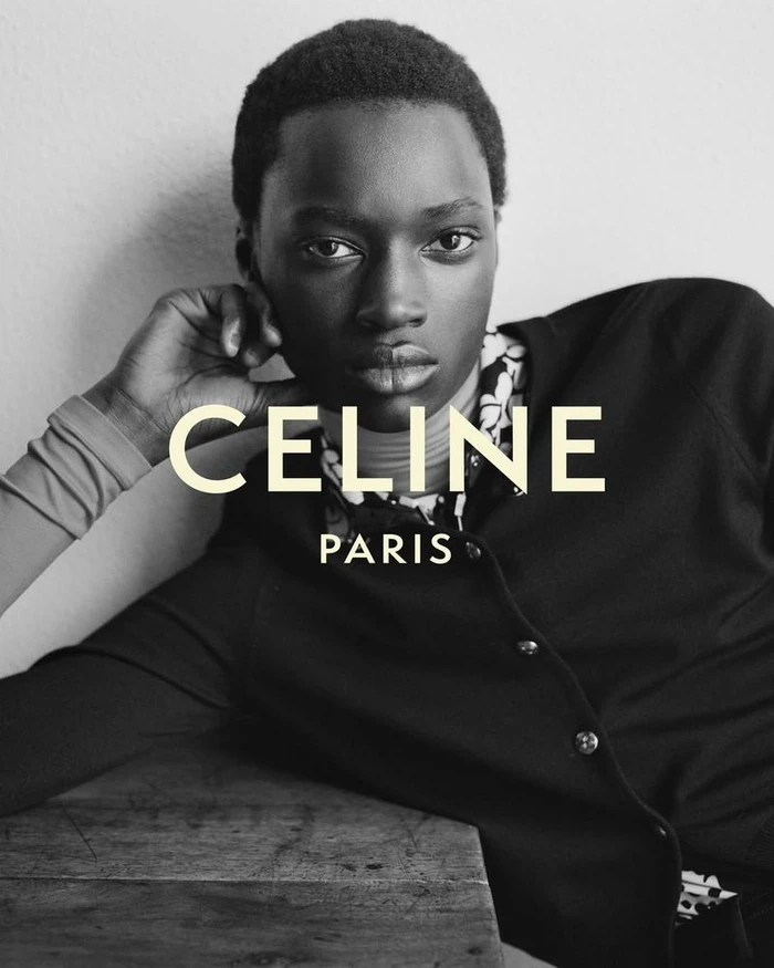 Black and white portrait of a Black male model in a dark buttoned cardigan resting his head on his hand for Celine.