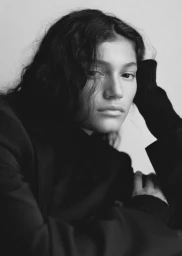 Close-up black and white portrait of a woman with dark wavy hair resting her head on her hand wearing a dark jacket.