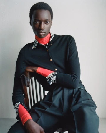 Model seated on a wooden chair wearing a layered black cardigan over a red turtleneck and pleated trousers against a neutral studio backdrop.