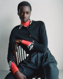 Model seated on a wooden chair wearing a layered black cardigan over a red turtleneck and pleated trousers against a neutral studio backdrop.