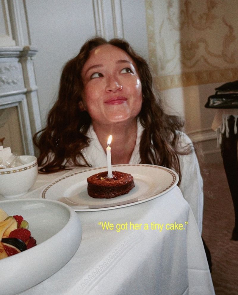 Young woman at a table with a tiny birthday cake with one candle, wearing a white shirt, indoors with natural light.