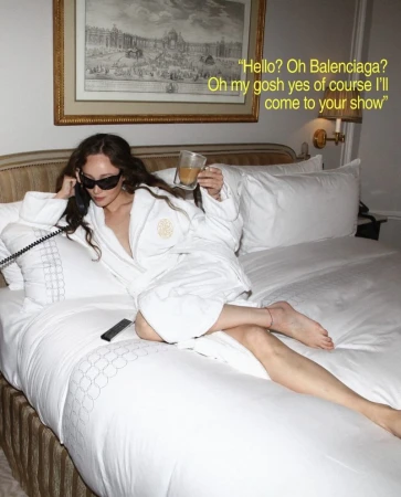 Adult female relaxing on a bed in a robe, holding a drink and a phone, with a framed picture on the wall in a hotel room.