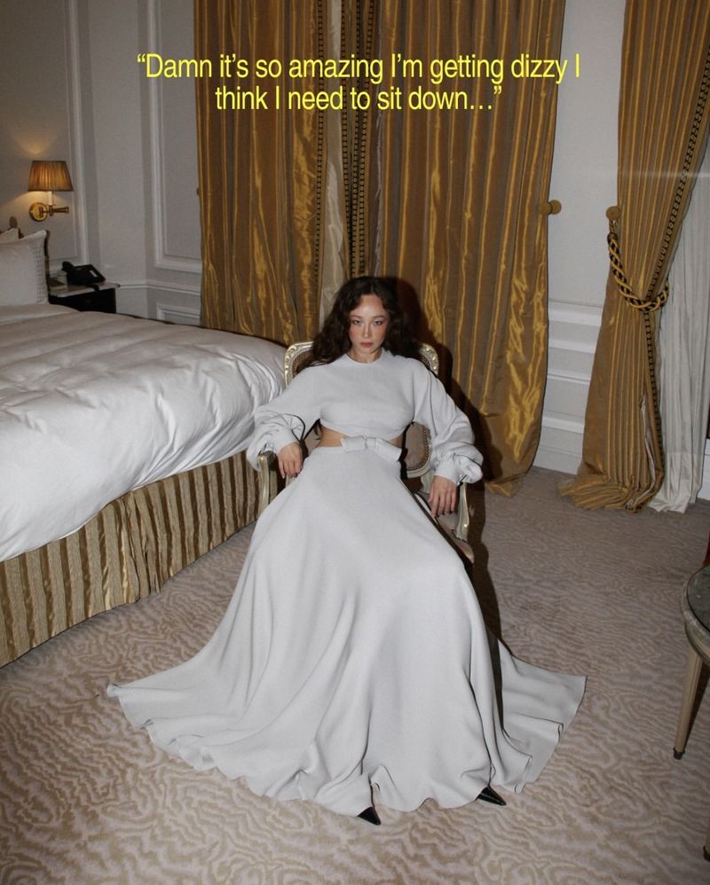 Woman with long curly hair sits in a long dress in an interior room with a bed, curtains, lamp, and patterned carpet.