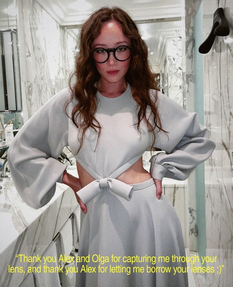 Woman in a gray dress and glasses stands in a bathroom with marble walls, looking at the camera with a neutral expression.