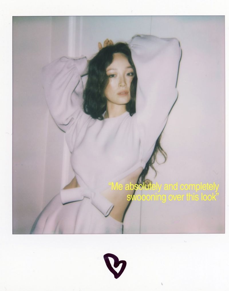 Portrait of an Asian woman in a light purple dress with her hands behind her head, with curly hair, posing in an indoor setting.