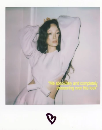 Portrait of an Asian woman in a light purple dress with her hands behind her head, with curly hair, posing in an indoor setting.