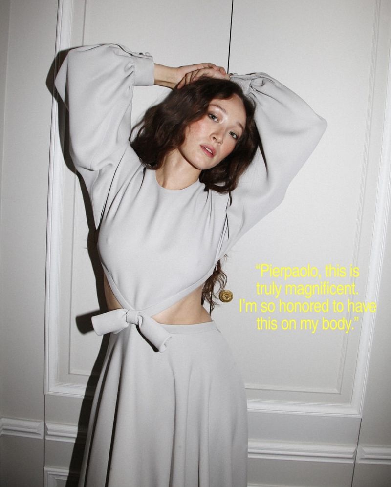 Havana Rose Liu posing with arms raised in a light grey cutout dress with balloon sleeves against a white paneled door.