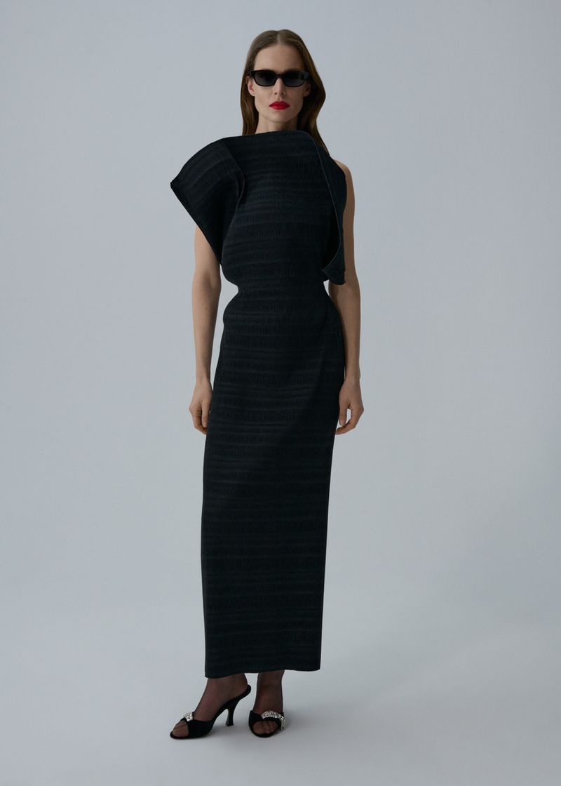 Elegant adult female poses in black sculptural dress and sunglasses in a studio, showcasing fashion and style in a full shot.