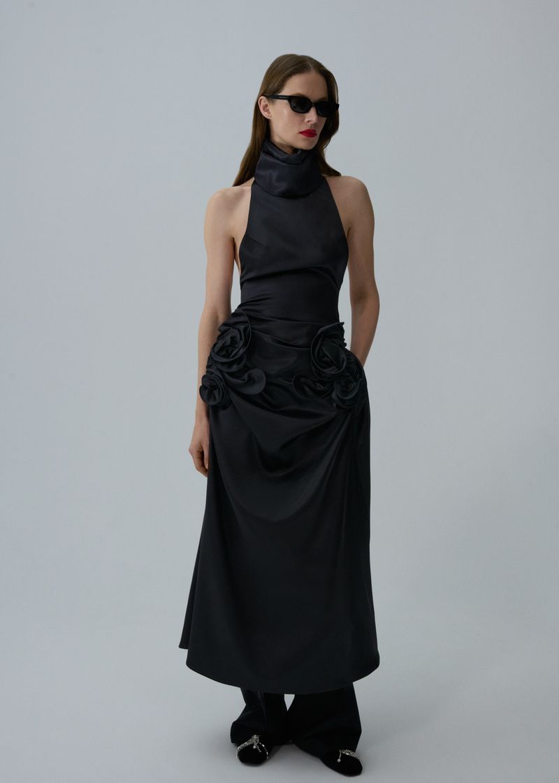 Elegant adult woman in a long black halter dress with floral details and sunglasses, posing in a studio setting against a white background.