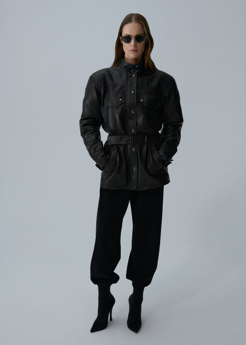 Woman in a belted black leather jacket, black pants, and stiletto heel boots stands in a full shot in front of a grey backdrop.