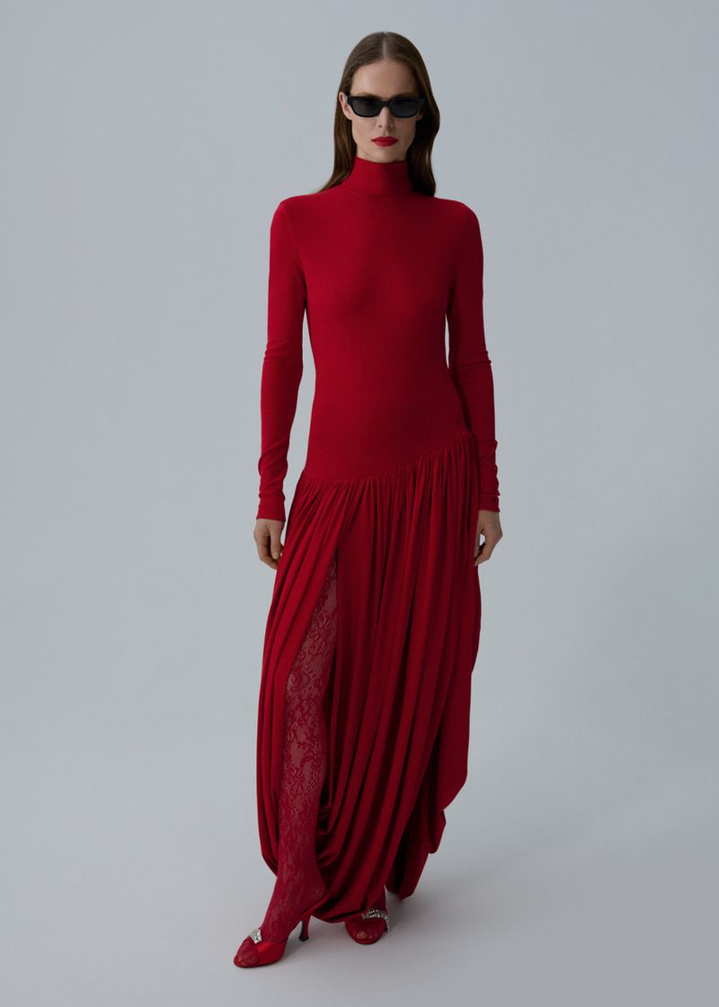 Woman in red turtleneck dress and sunglasses standing in studio. Full-length fashion shot against plain light-grey background.