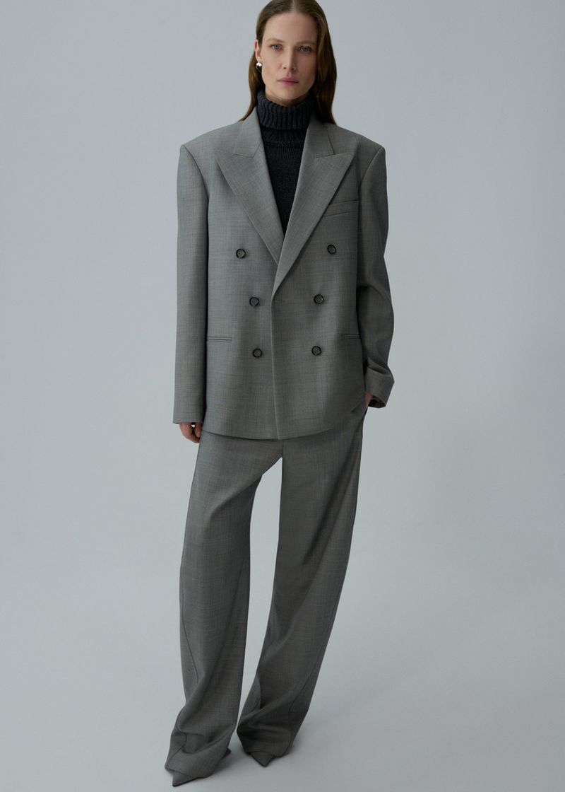 Adult female wearing gray suit with black turtleneck sweater in a full-length studio shot with soft lighting against a neutral backdrop.