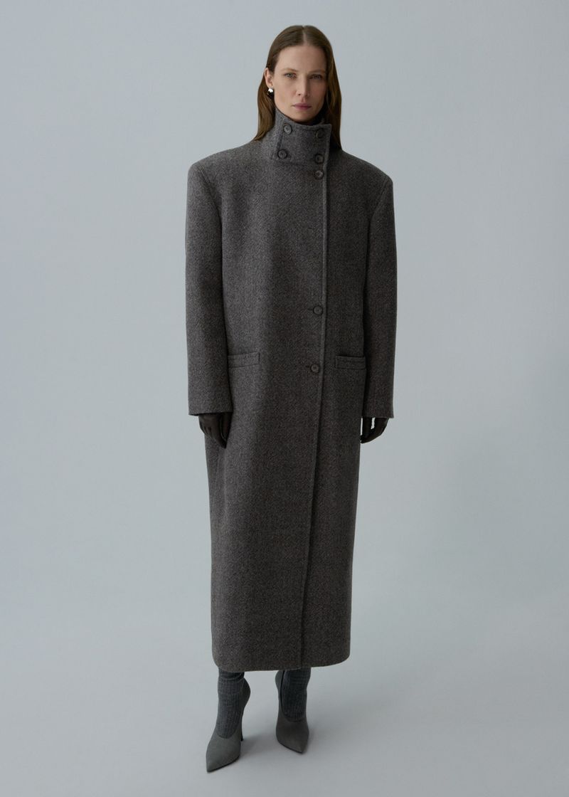 Full-length shot of an adult female model in a long grey wool coat, gloves, and grey heels, standing against a pale grey backdrop.