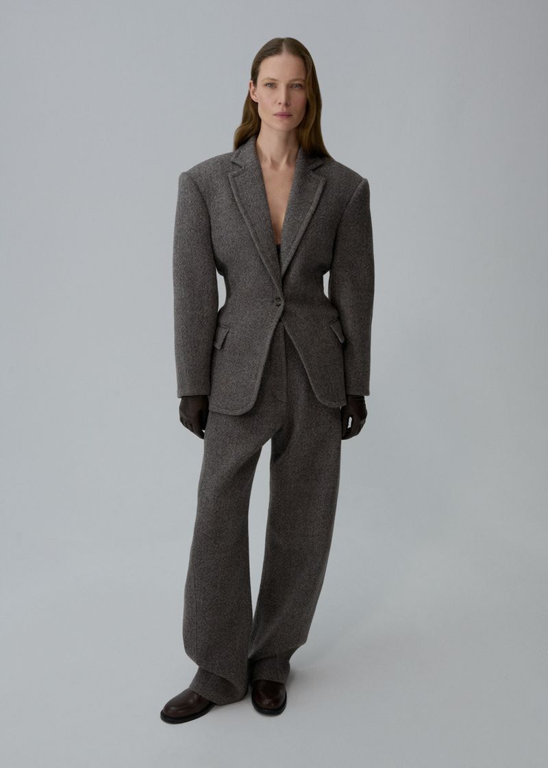 Full-length shot of a woman with brown hair wearing a grey wool blazer and slacks, with brown gloves and shoes against a grey backdrop.