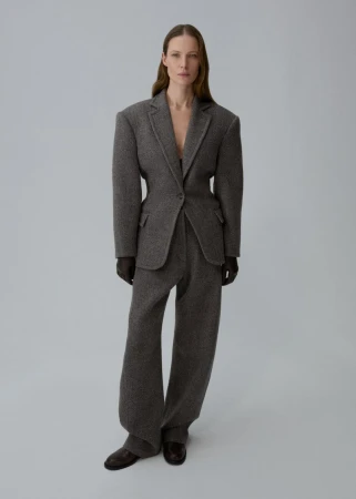 Full-length shot of a woman with brown hair wearing a grey wool blazer and slacks, with brown gloves and shoes against a grey backdrop.