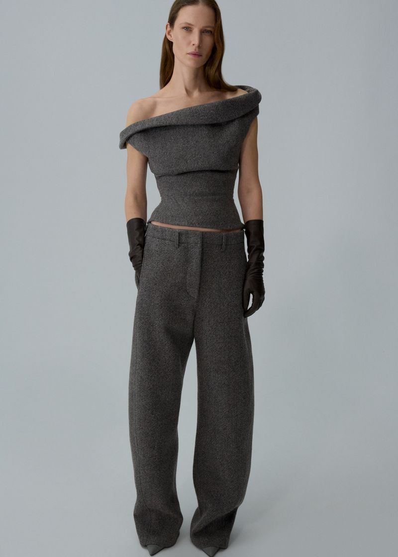 Indoor shot of a woman in a gray, off-the-shoulder top and wide-leg pants, complemented by leather gloves, against a light gray background.