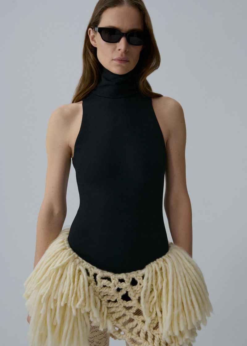 Model wearing a black sleeveless turtleneck bodysuit, dark sunglasses, and cream fringe knit skirt against a studio backdrop.