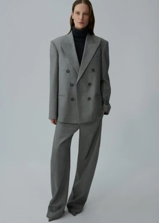 Model Angelika Wierzbicka poses in an oversized grey double-breasted suit and black turtleneck against a neutral grey studio backdrop.