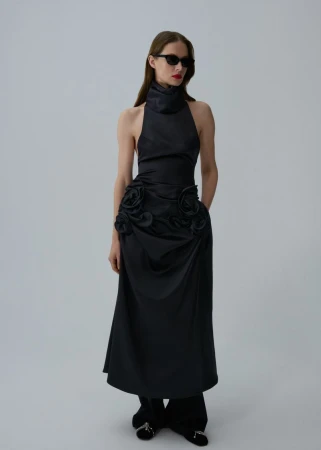 Model Angelika Wierzbicka wearing a black high-neck rosette maxi dress, black trousers, and sunglasses against a grey studio backdrop.