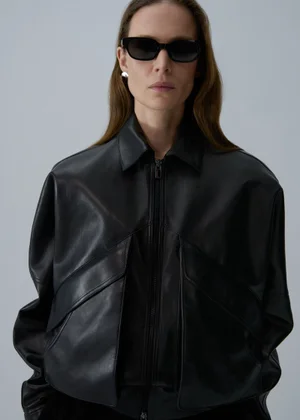 Model wearing a black leather jacket and sunglasses against a plain grey studio backdrop for Magda Butrym