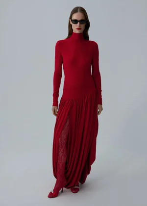 Model in dark sunglasses wearing a long red turtleneck dress and lace tights against a grey studio backdrop