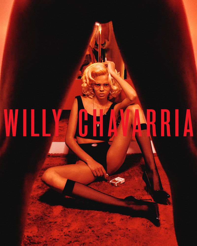 A blonde woman sits on a carpeted floor in black clothing, hosiery and heels with a pack of cigarettes next to her, with 'Willy Chavarria' written across the scene.