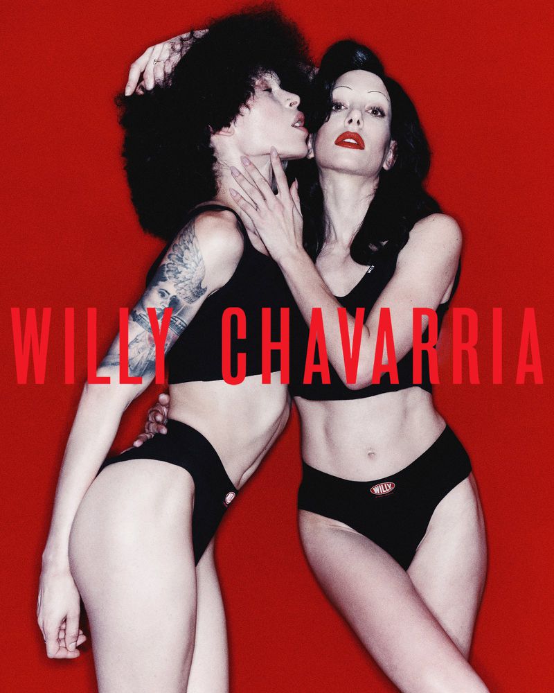 Two adult females pose intimately in front of a red background. They wear black tops and underwear. The brand name 'Willy Chavarria' is visible.