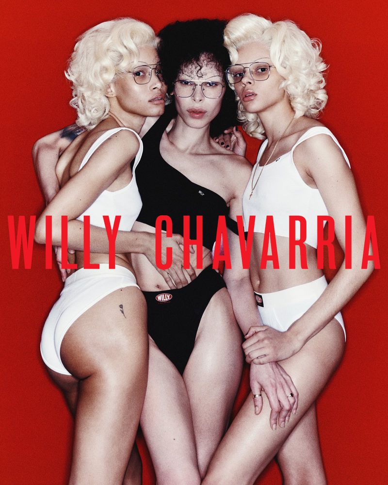 Three models in Willy Chavarria branded swimwear pose closely against a vibrant red backdrop, showcasing a blend of fashion and style.