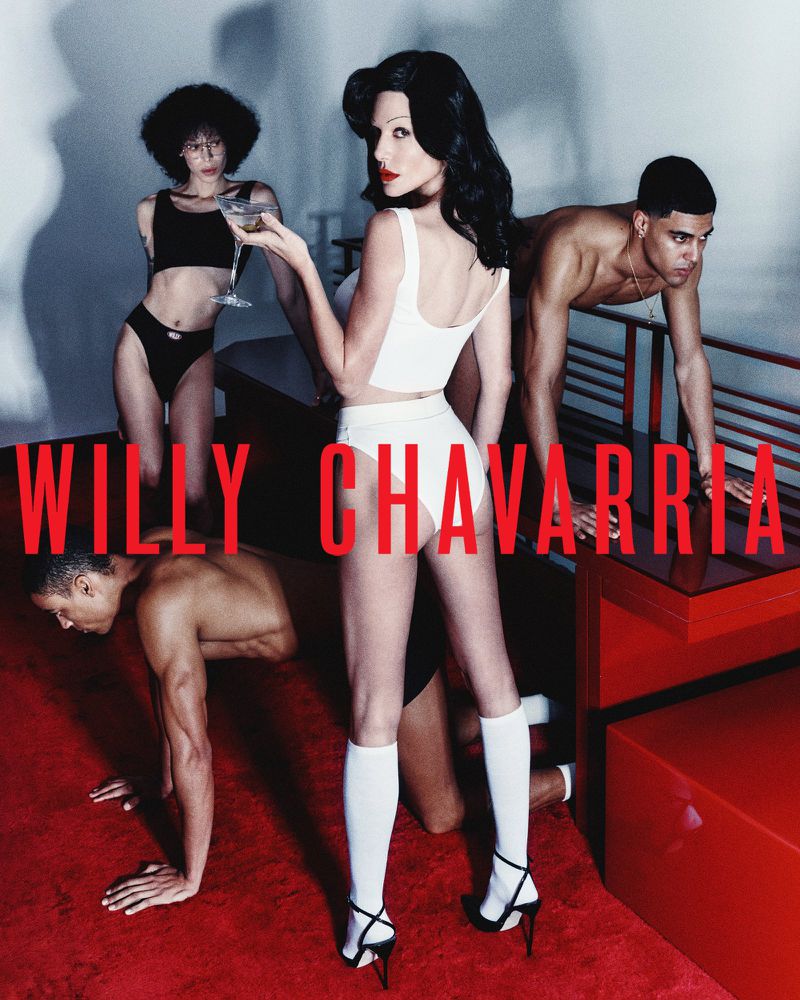 A group of fashion models pose in a photo studio wearing white underwear-style garments, hosiery and heels, accompanied by shirtless male models.