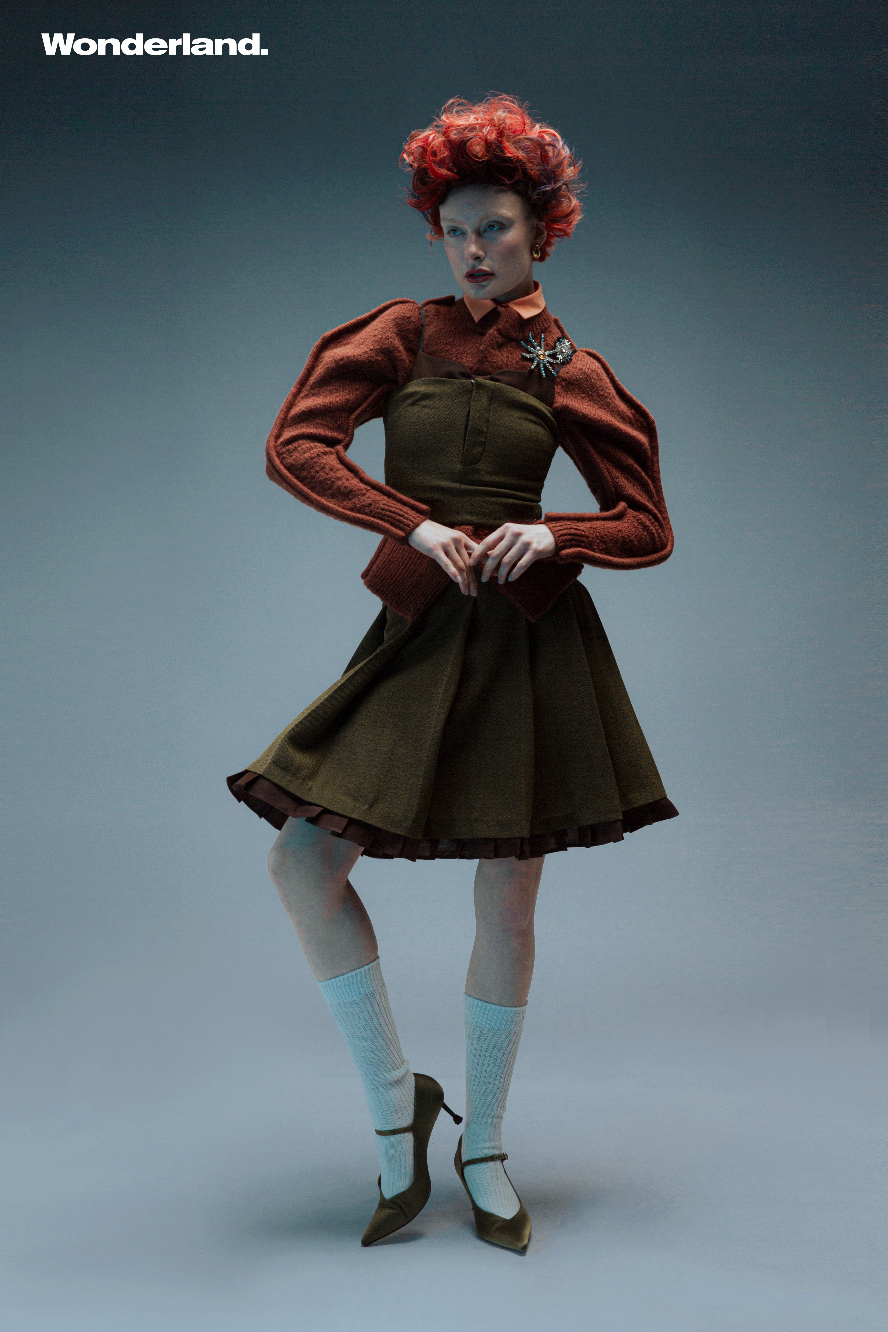 Fashion editorial shot of a redhead woman in a brown and olive green outfit, posing in a studio with a light blue-grey background.