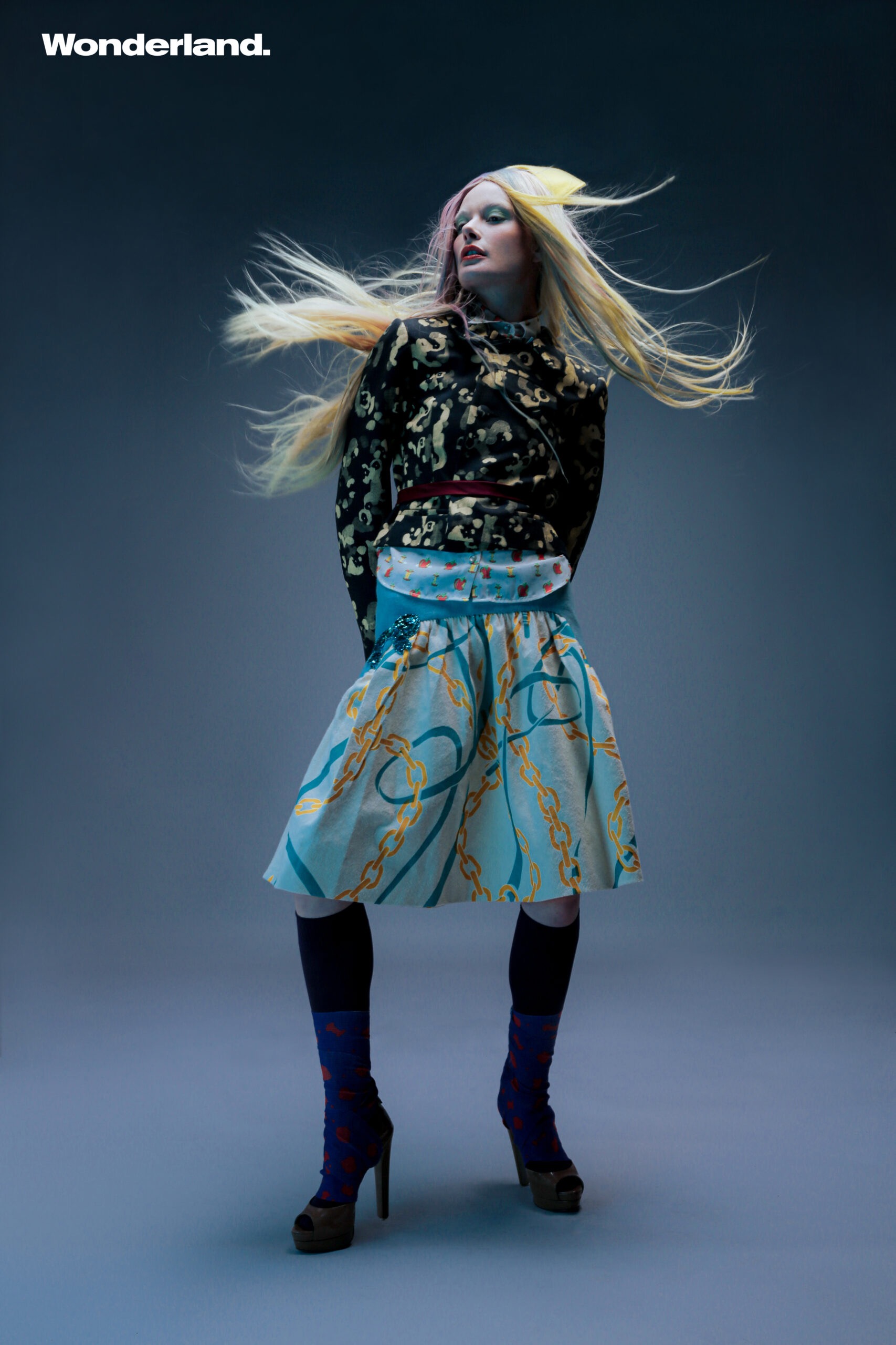 Fashion editorial shot of a woman with blonde hair wearing a printed top and skirt, standing in front of a grey background with logo.