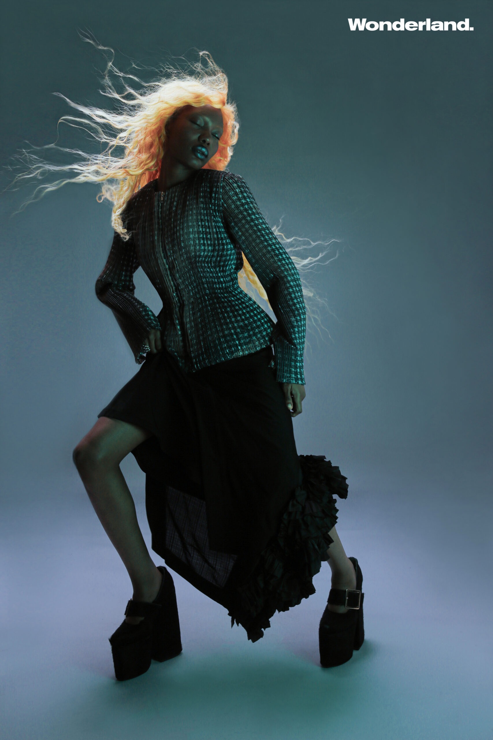 Fashion editorial shot of a woman with blonde hair wearing an embellished jacket, black skirt, and platform shoes, posing dynamically.