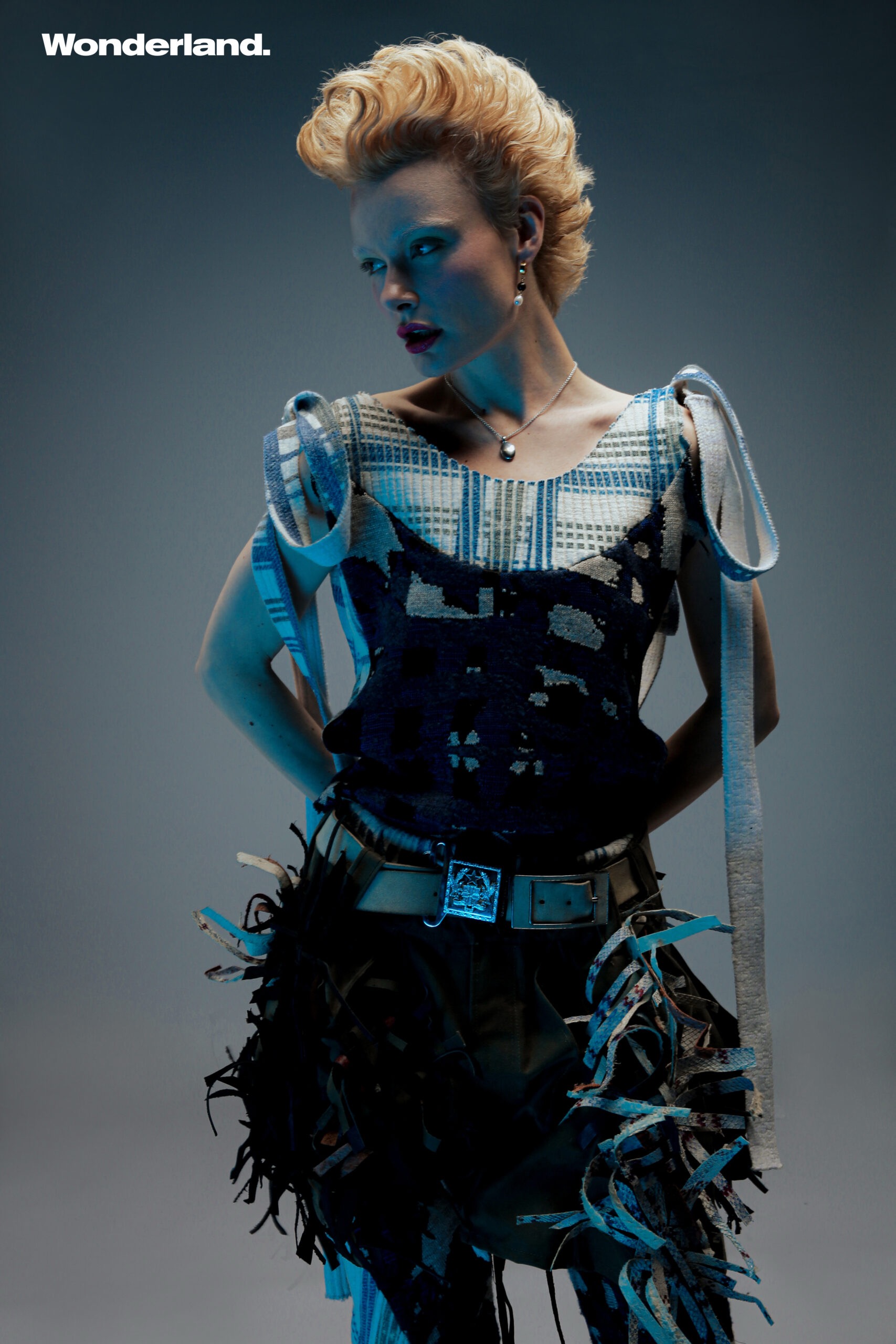Blonde woman in stylish textured top and fringed belt poses in a fashion shot with cool lighting and a grey studio backdrop.