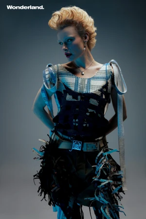 Blonde woman in stylish textured top and fringed belt poses in a fashion shot with cool lighting and a grey studio backdrop.