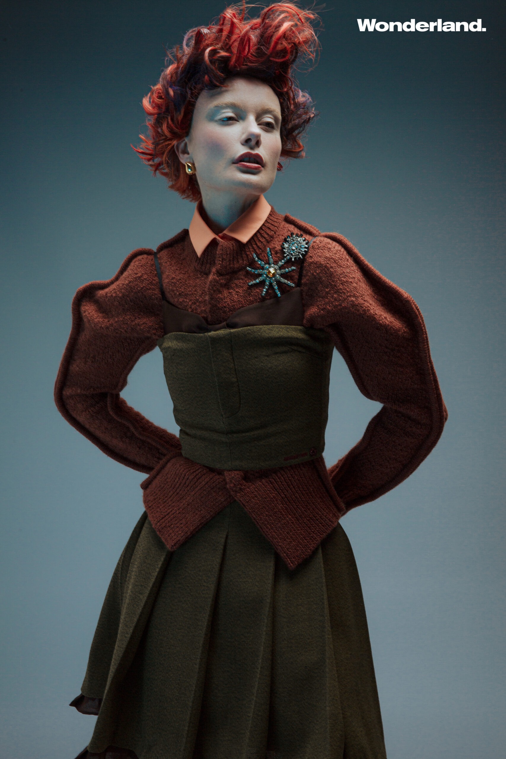 Fashion editorial shot featuring a posed woman with red hair wearing an ornate brown outfit with a green dress and an insect brooch.