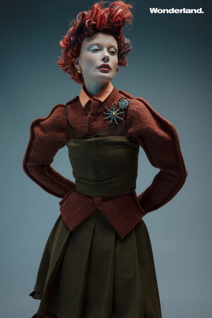 Fashion editorial shot featuring a posed woman with red hair wearing an ornate brown outfit with a green dress and an insect brooch.