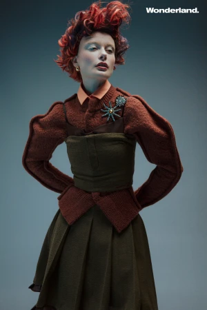 Fashion editorial shot featuring a posed woman with red hair wearing an ornate brown outfit with a green dress and an insect brooch.