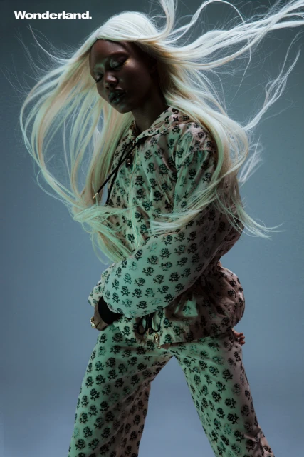 A woman with platinum blonde hair poses in patterned outfit, eyes closed, against a gradient blue background, showcasing a fashion editorial shot.