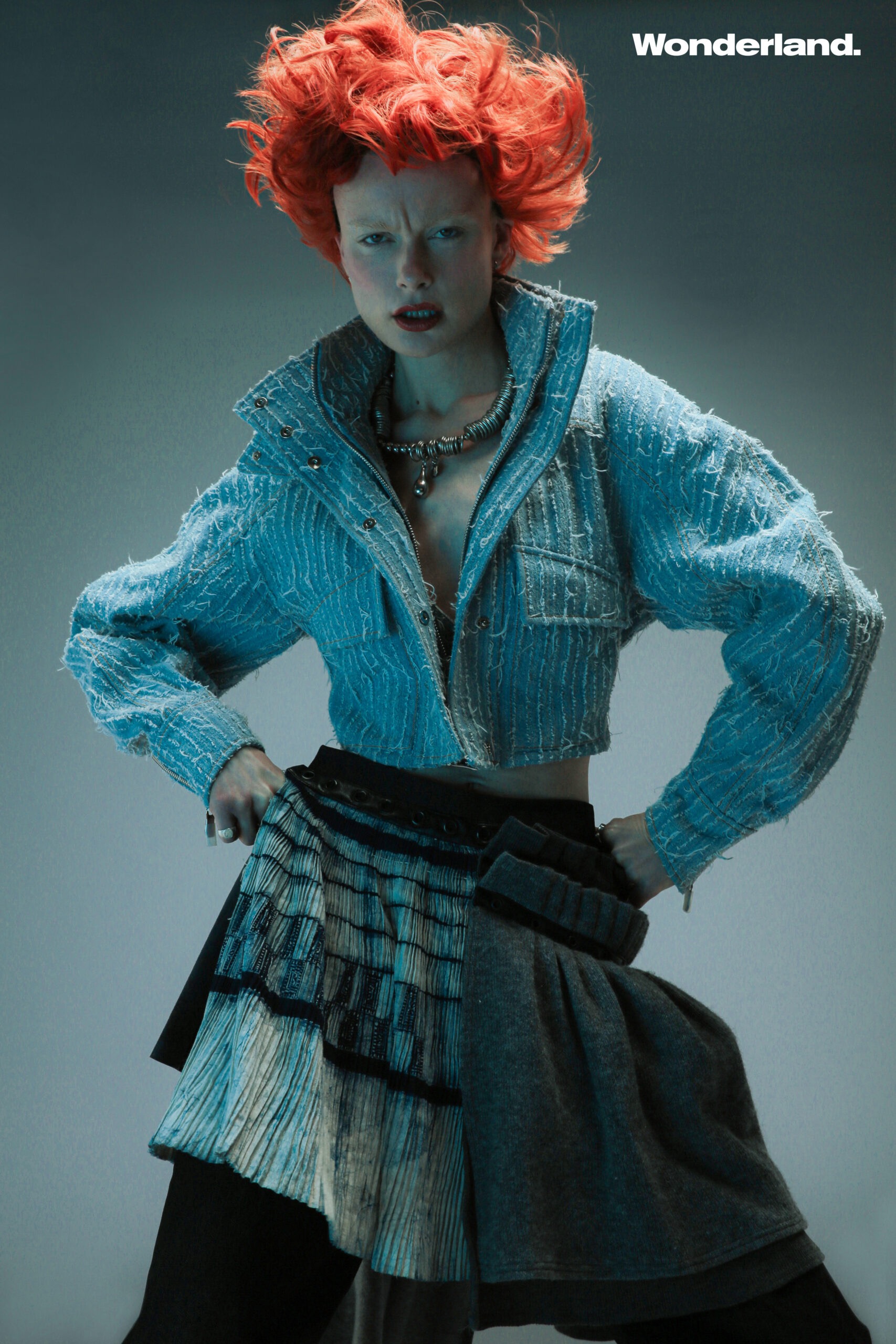 An adult female fashion model with bright red hair is posing in a studio wearing a short denim jacket and patchwork skirt for Wonderland.