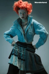Model with vibrant red hair wearing a cropped light blue textured jacket and layered skirt against a grey studio backdrop.