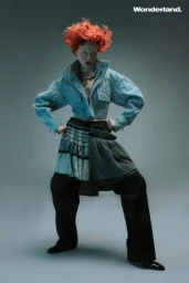 Fashion model with voluminous orange hair wearing a cropped light blue jacket, layered skirt, and wide-leg trousers against a grey backdrop.