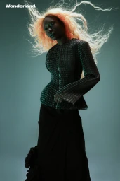 Black woman with wild flowing blonde-tinted hair wearing a textured patterned top against a muted teal studio backdrop.