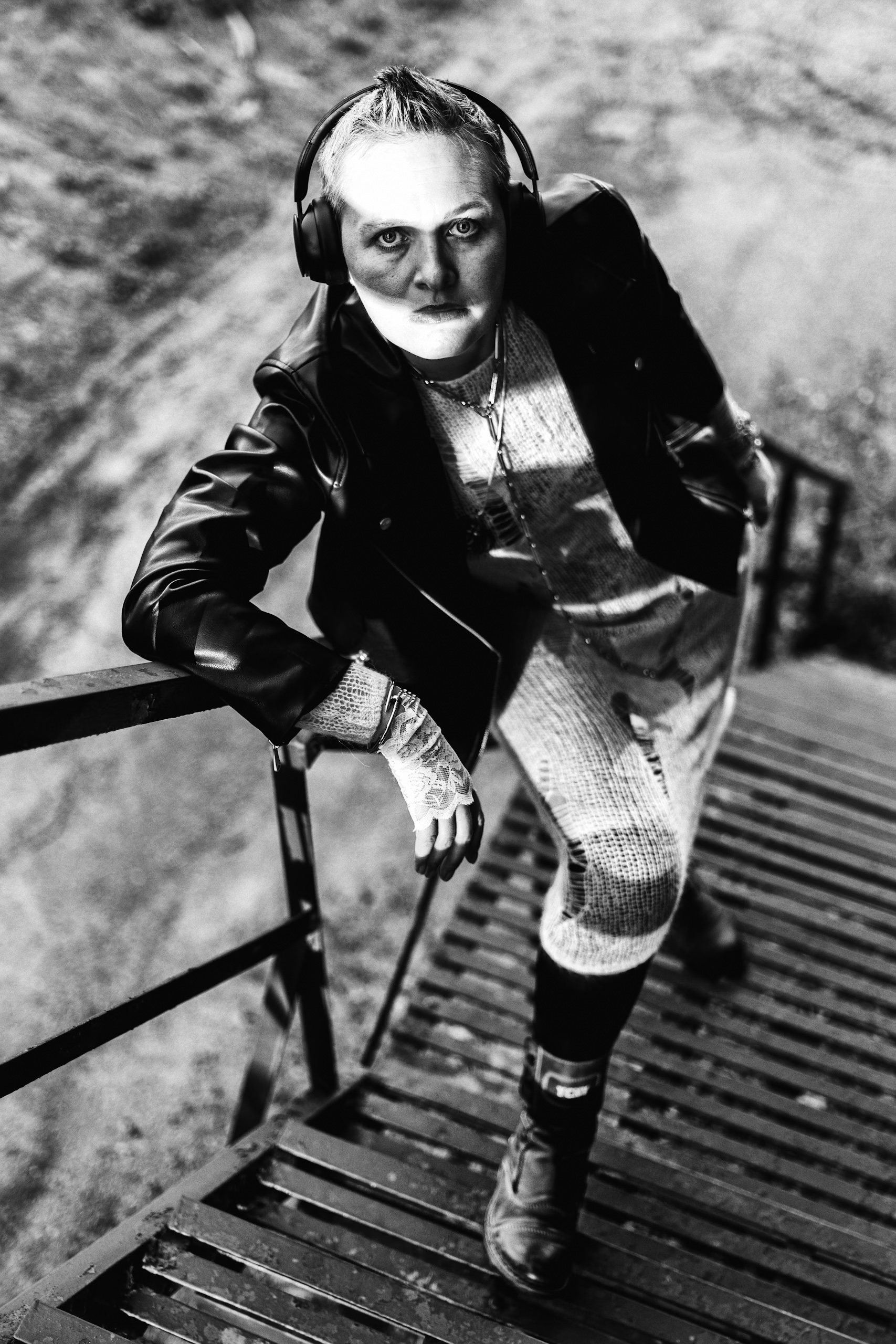 Black and white portrait of a woman wearing headphones and a jacket, leaning on a metal railing outdoors, with a high-angle perspective.