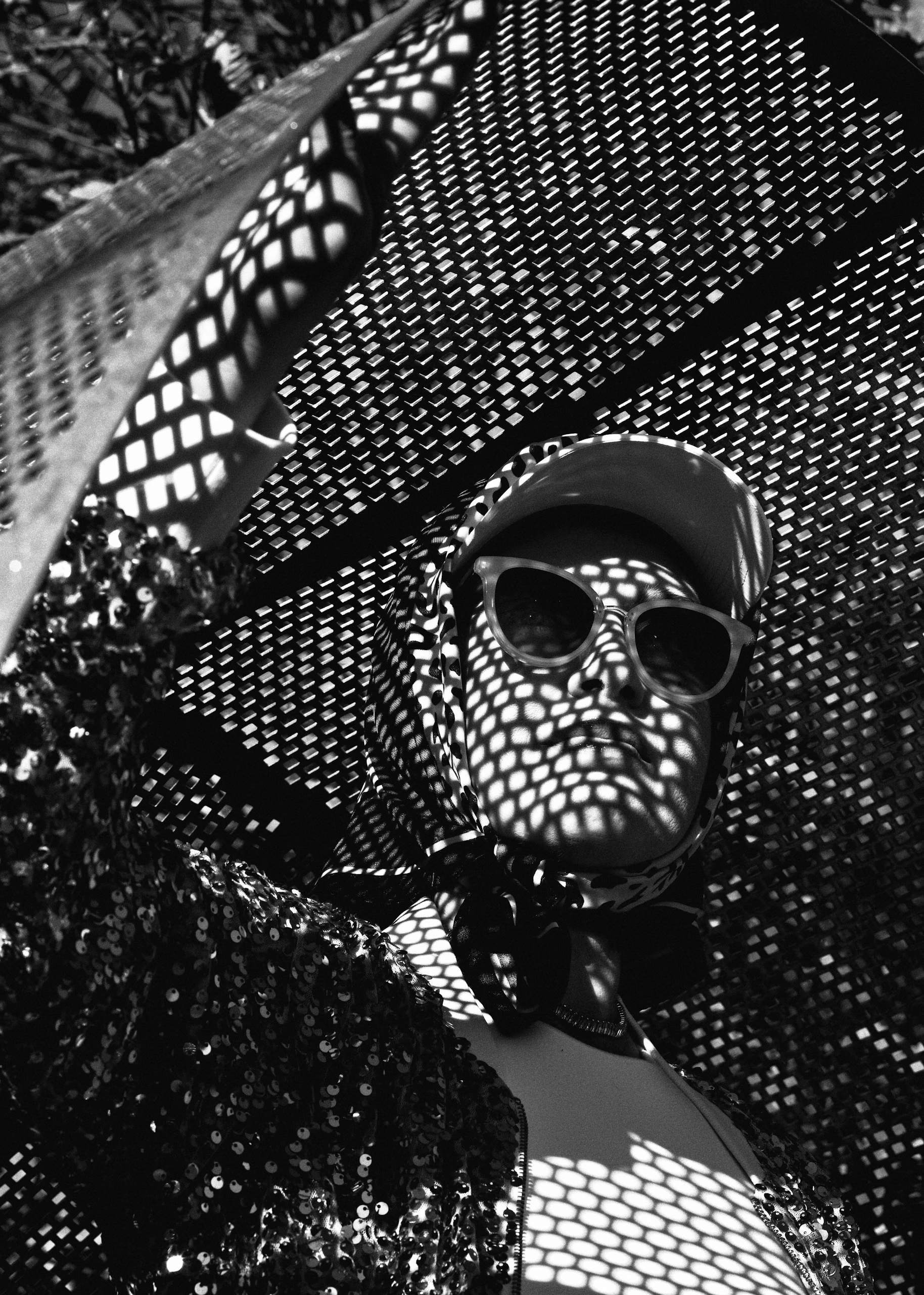 Black-and-white portrait of an adult wearing sunglasses and a cap, patterned with light from a perforated metal structure, outdoors.