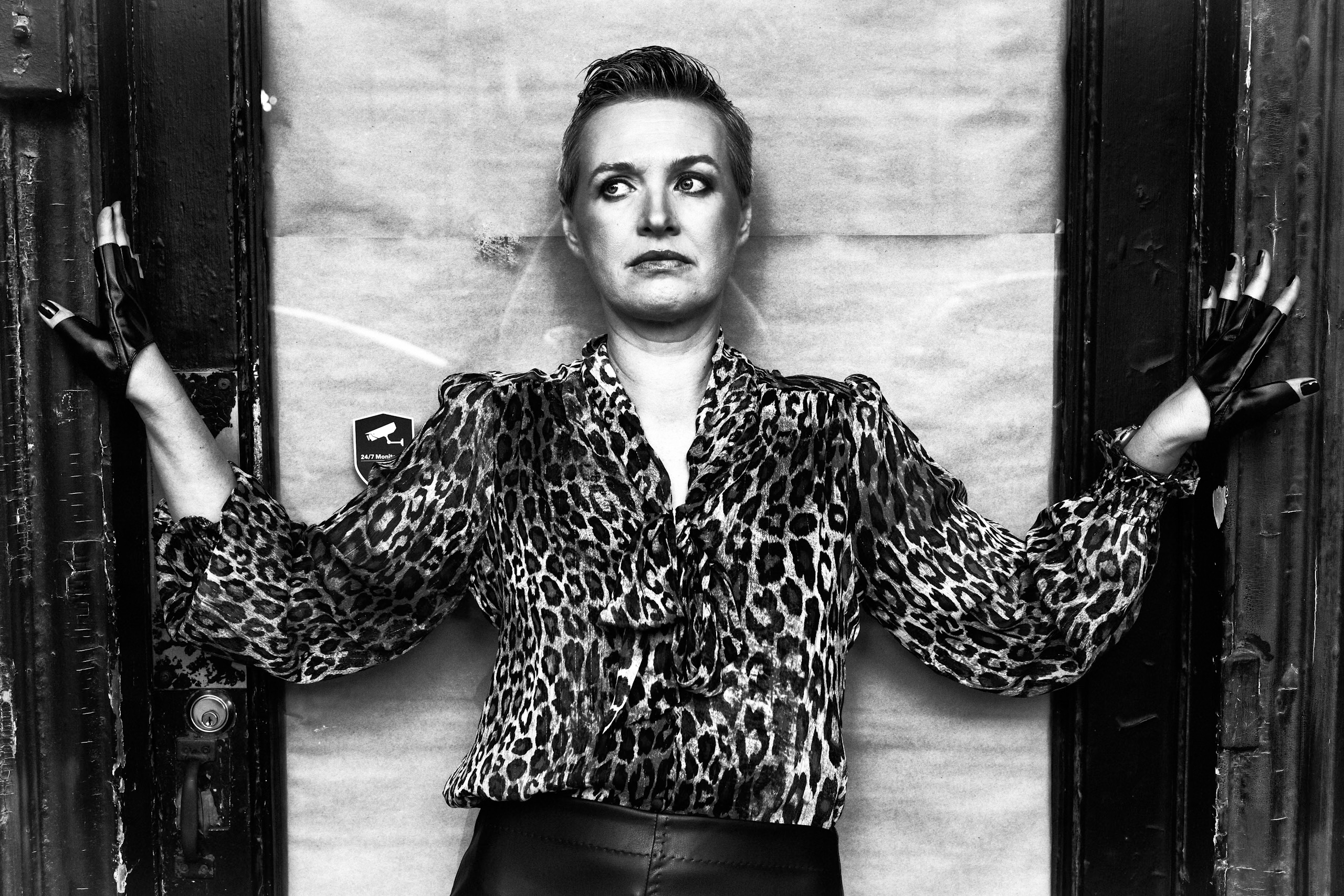 Black and white portrait of a woman in a leopard print blouse standing in a doorway with extended arms wearing gloves.